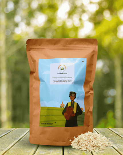 The Deep Soil Pahadi Brown Rice package with a small pile of loose unpolished grains on a wooden table and sunlit forest background