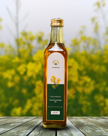 The Deep Soil Cold-Pressed Yellow Mustard Oil 500ml glass bottle on a rustic wooden table with a blurred field of yellow mustard flowers